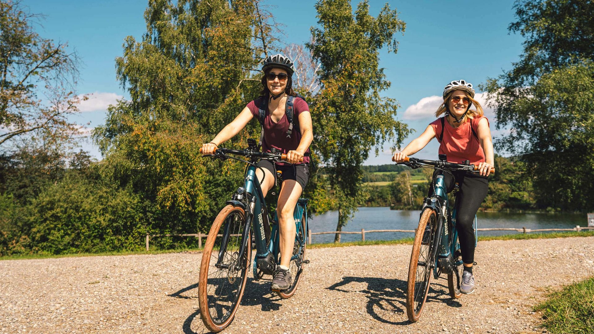 Two women cycling on a trail by a lake on a sunny day