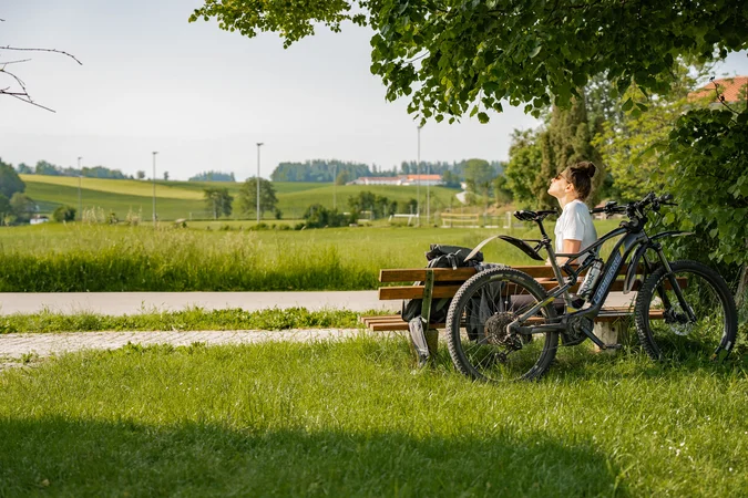 Frau auf Parkbank neben Fahrrad genießt sonnige Landschaft