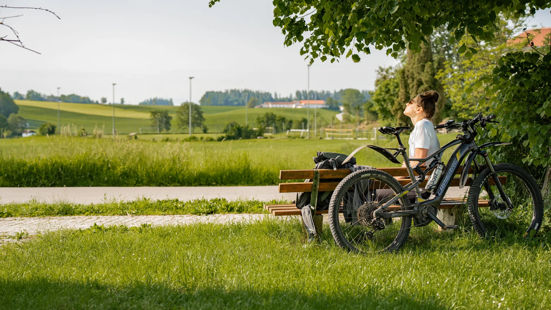 Frau auf Parkbank neben Fahrrad genießt sonnige Landschaft