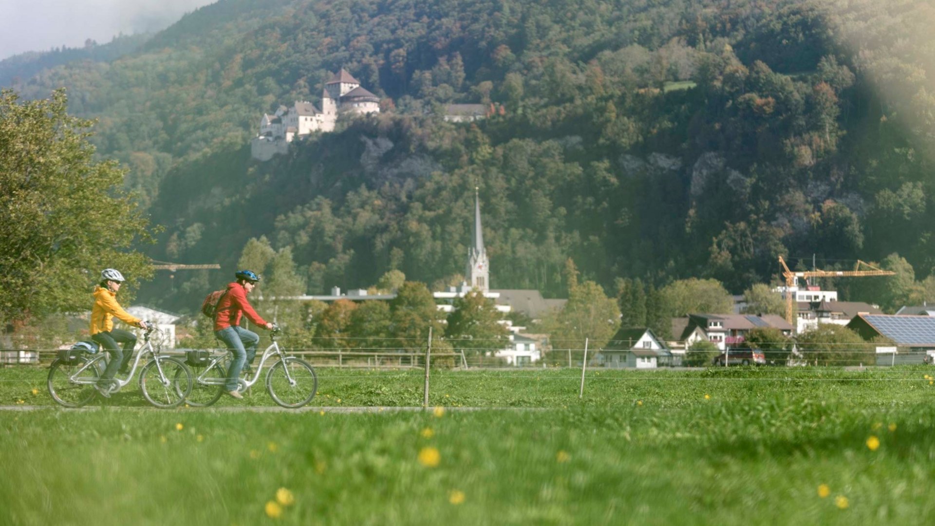 Two cyclists cycling with mountains, castle and village in the background