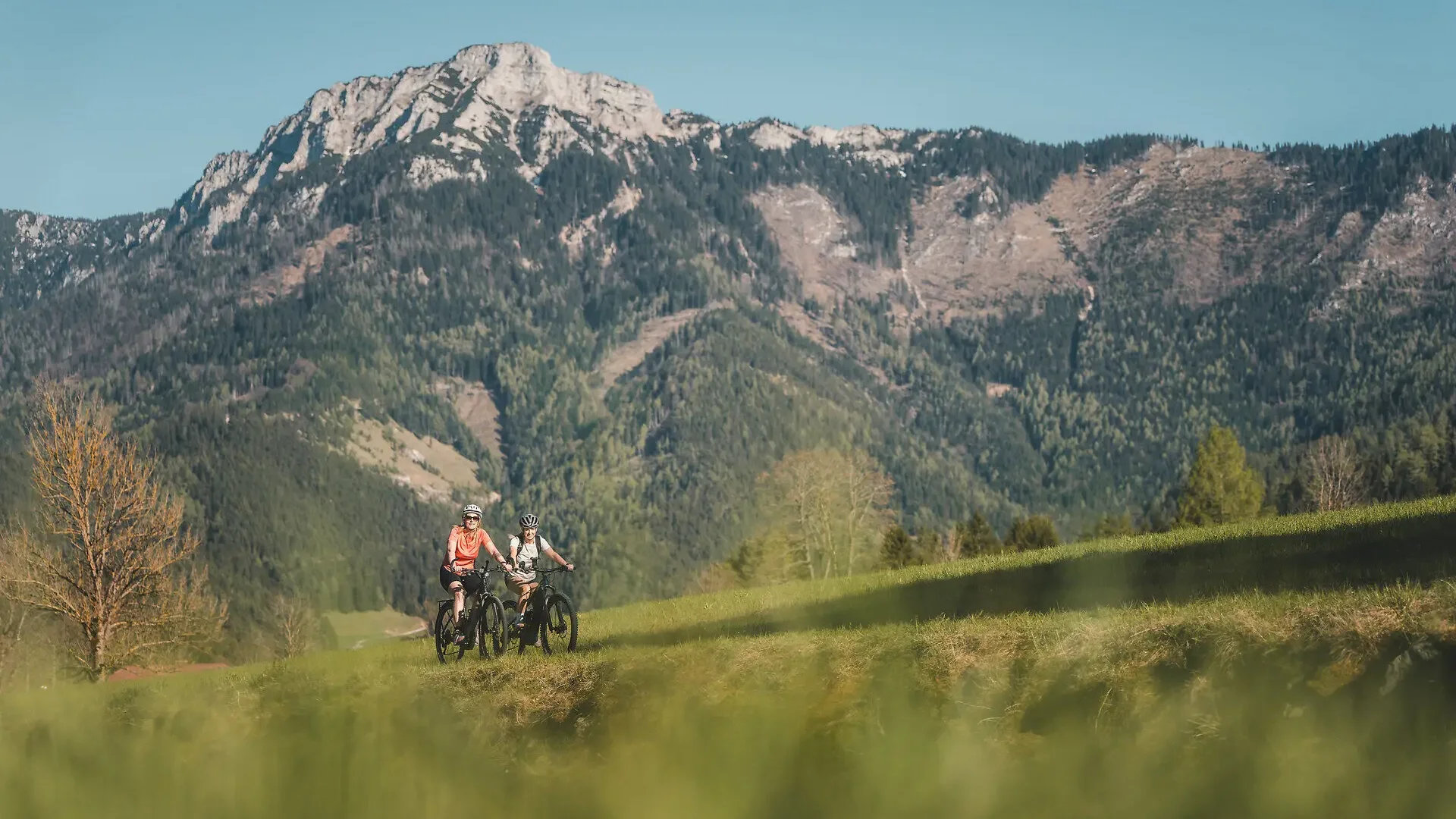 Tour-Detail Zwei Radfahrer auf grünem Hügel vor großen Bergen unter blauem Himmel