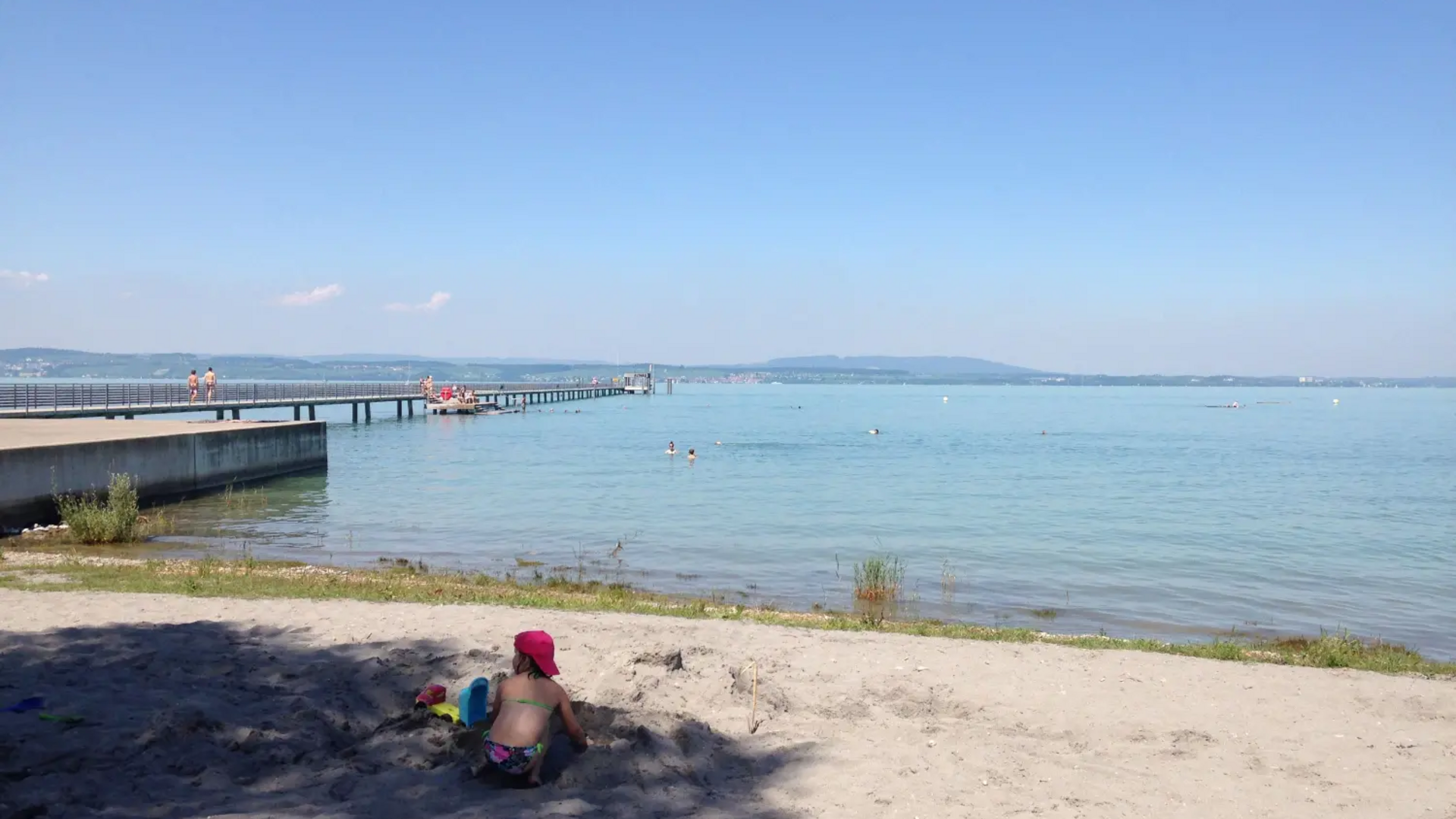Child playing on sandy beach near lake with pier and swimmers
