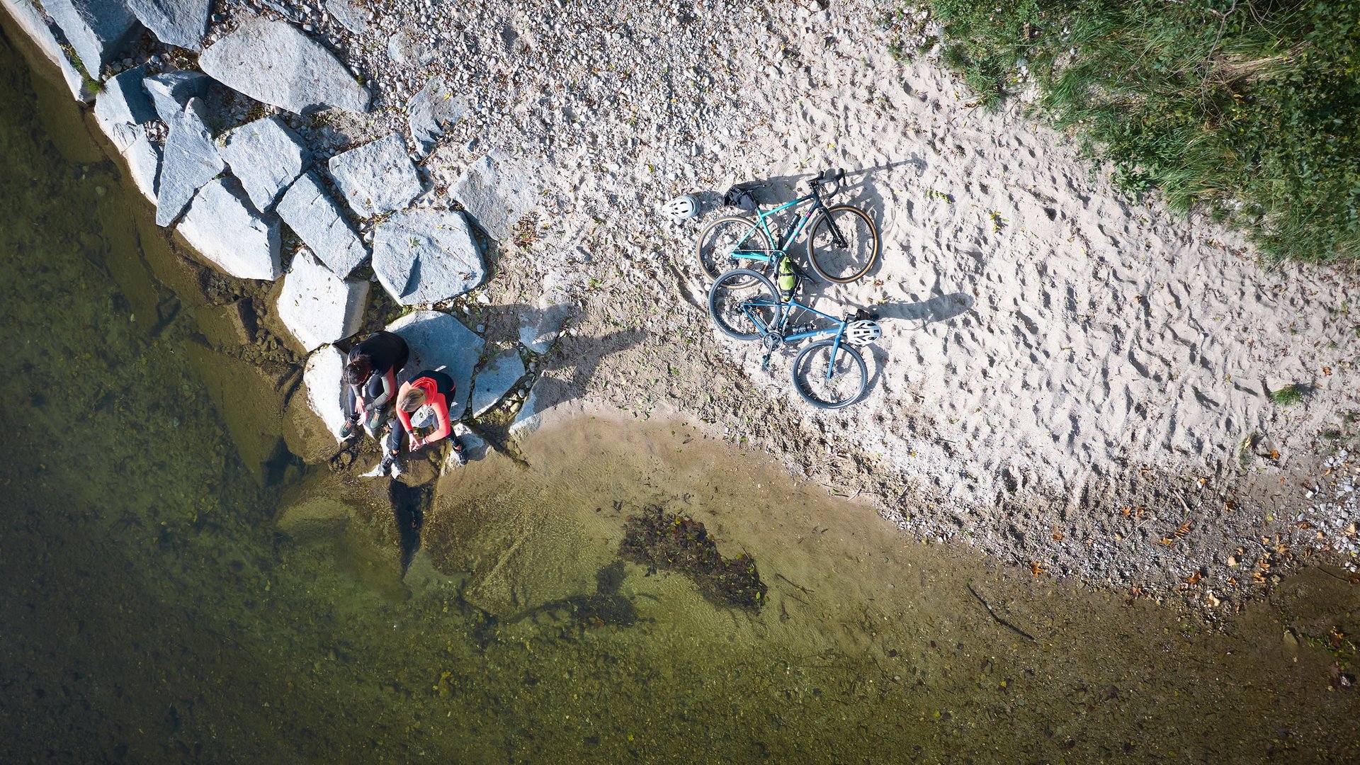 Two people sitting by shore with two bikes on sand and rocks