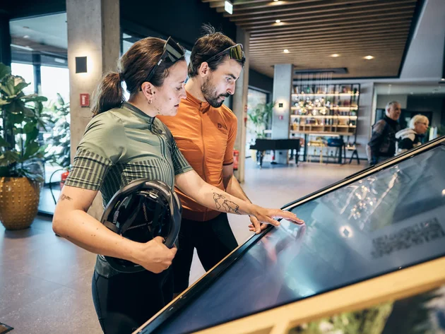 Zwei Radfahrer schauen auf einen großen Touchscreen in einem modernen Innenraum