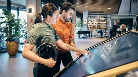 Zwei Radfahrer schauen auf einen großen Touchscreen in einem modernen Innenraum