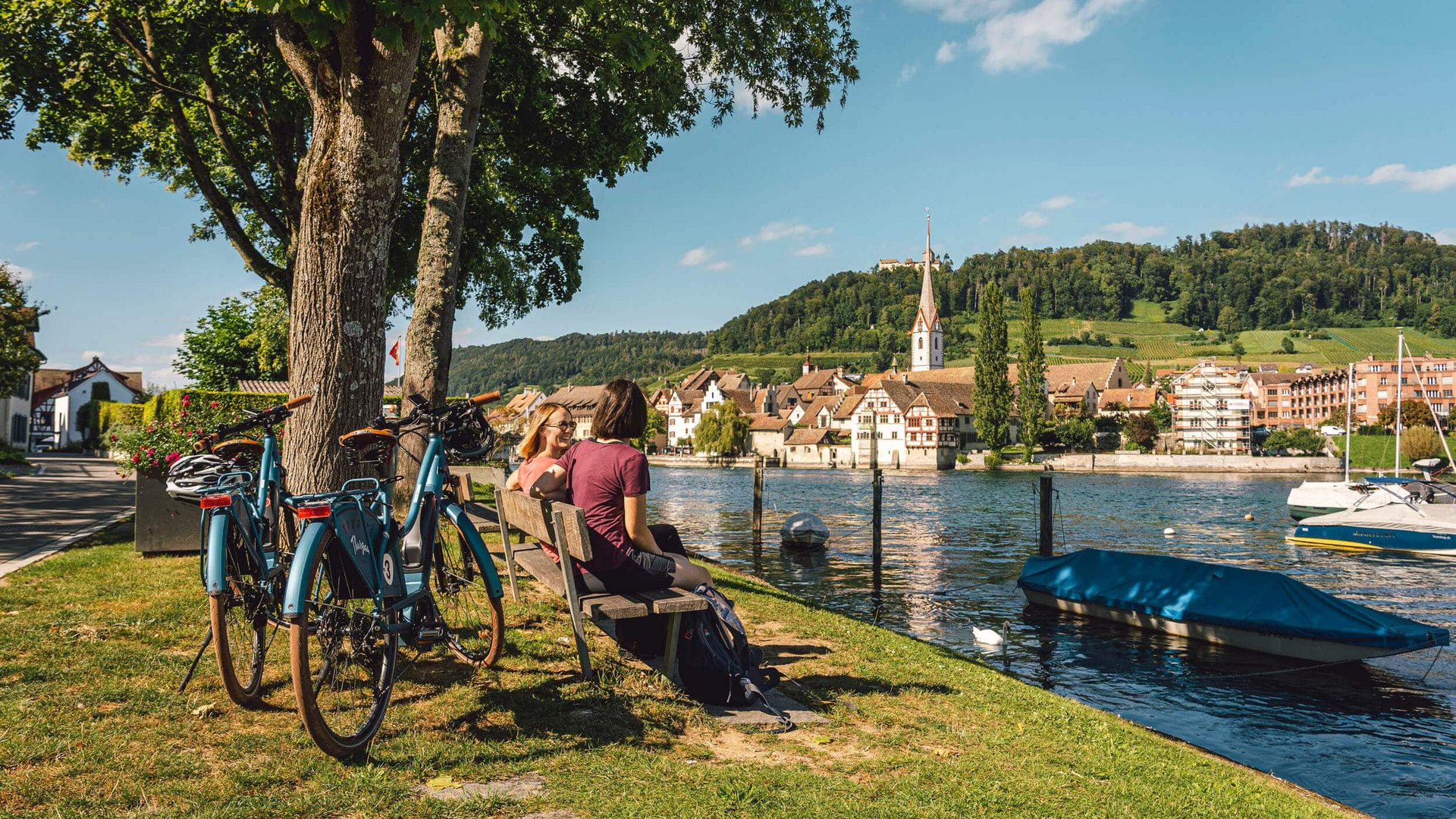 Two people sitting on a bench by the river with bikes and village with hills in background