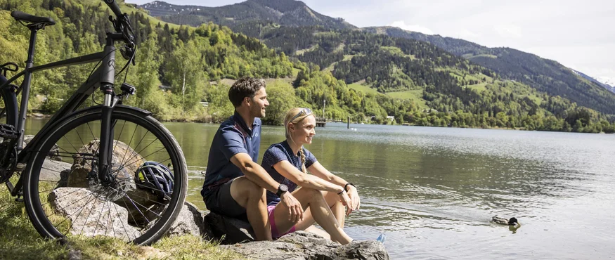 5 Gründe, warum Maishofen der Geheimtipp für Genussradler ist © Velontour Paar sitzt am Seeufer mit Fahrrad und Blick auf Berge
