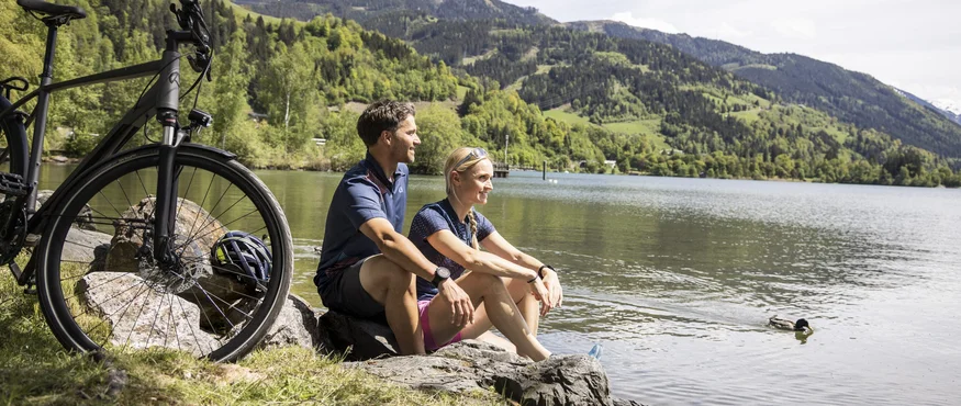 Paar sitzt am Seeufer mit Fahrrad und Blick auf Berge