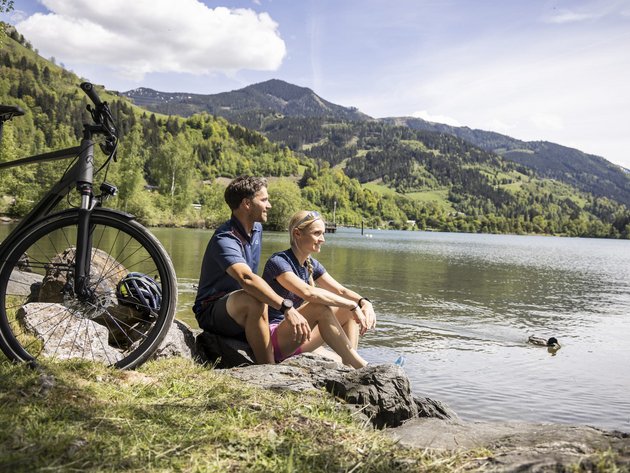 Paar sitzt am Seeufer mit Fahrrad und Blick auf Berge