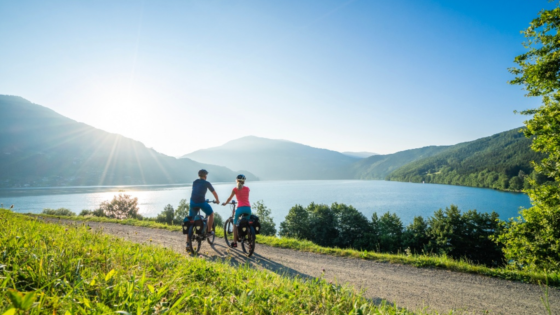 Two cyclists on path by lake with mountains and sun