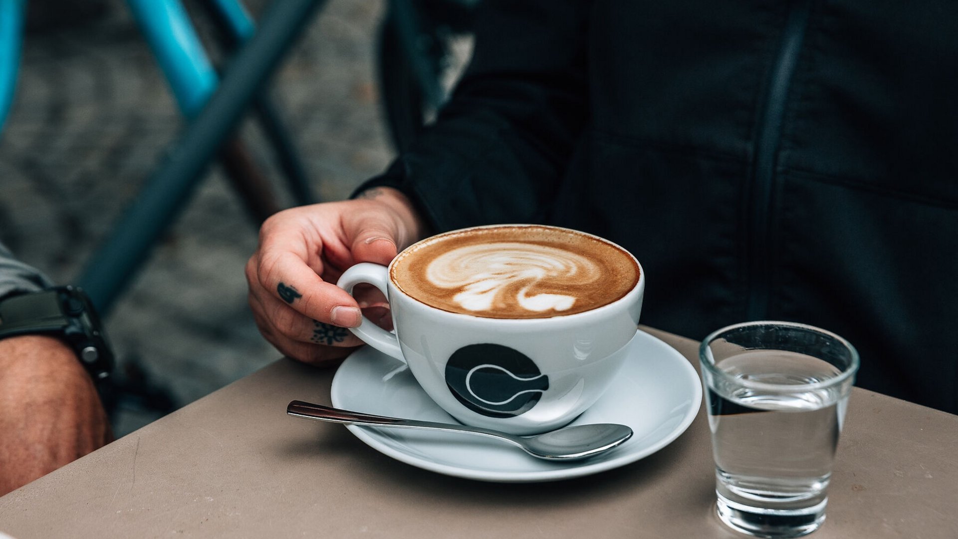 Hand holding cappuccino cup with foam art, glass of water, and spoon on table