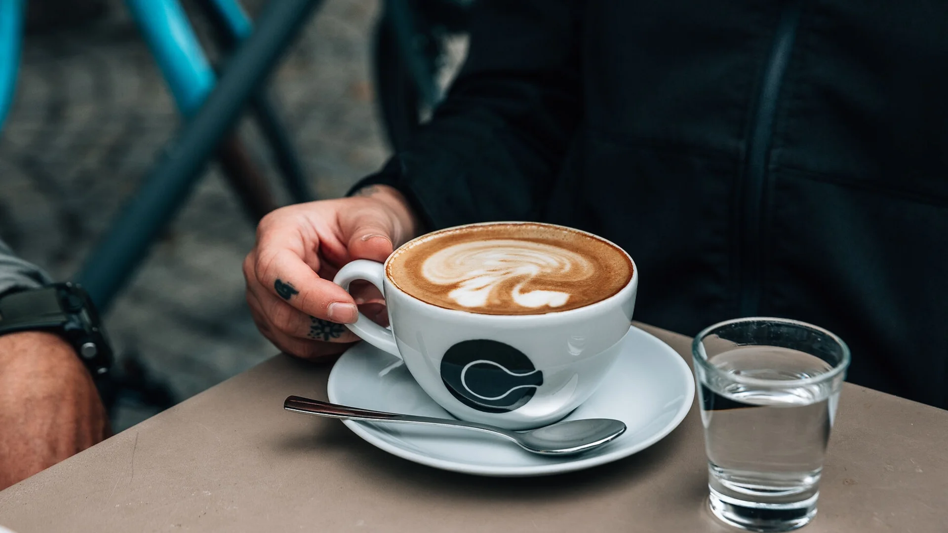 Hand hält Cappuccino-Tasse mit Milchschaum, Glas Wasser und Löffel auf dem Tisch