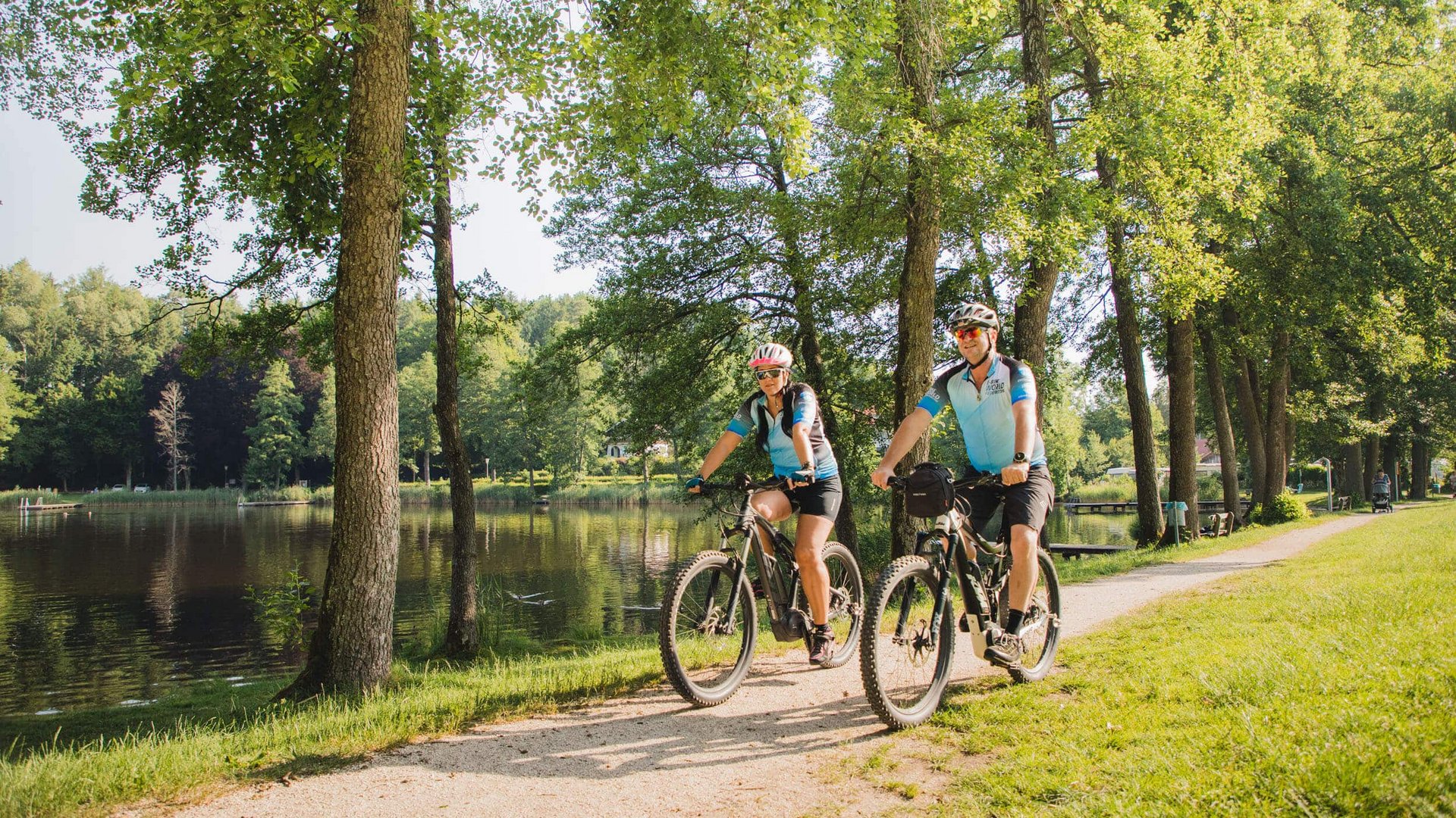 Two cyclists riding on a forest path beside a lake on a sunny day