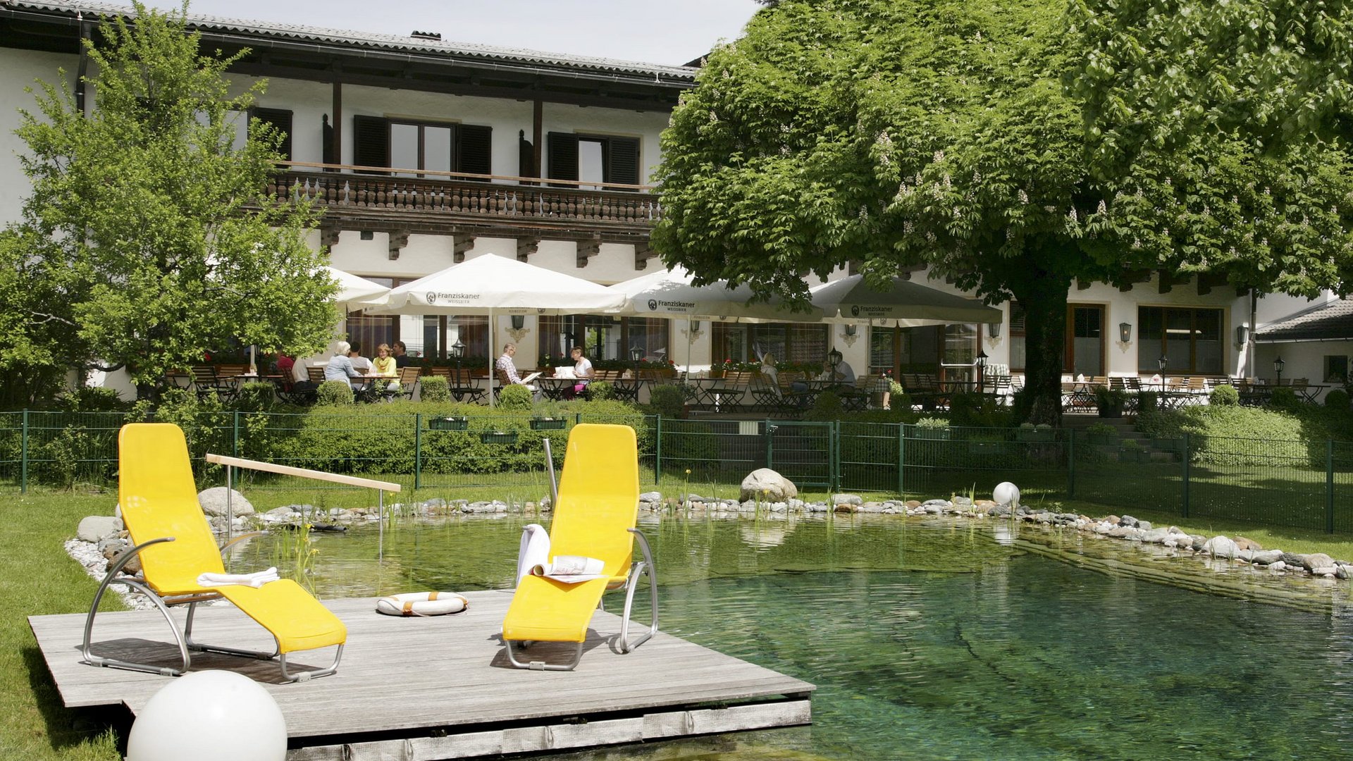 Sunny terrace with yellow lounge chairs beside a clear pond in front of a restaurant