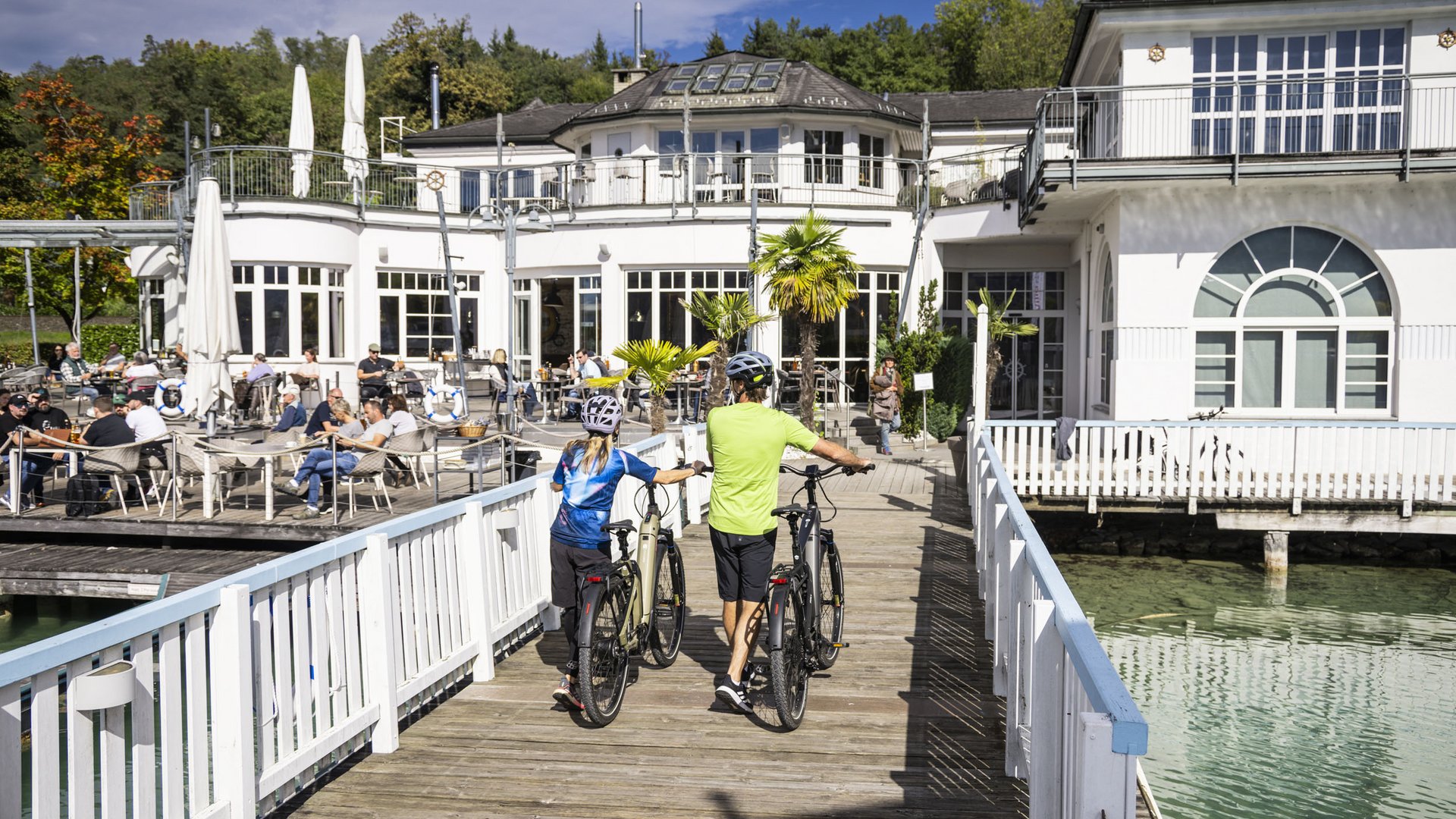 Two cyclists walking bikes on pier by a lakeside café on a sunny day