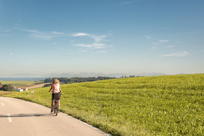 Frau fährt Fahrrad auf Landstraße neben grünem Hügel bei klarem Himmel