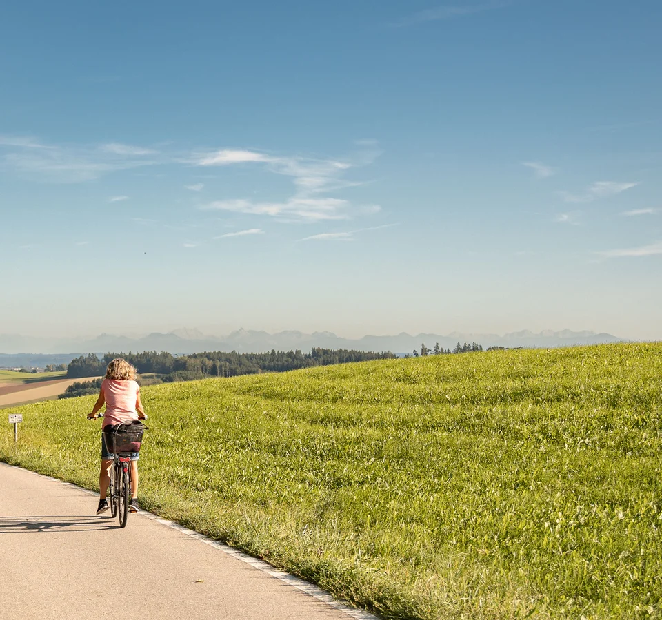 Frau fährt Fahrrad auf Landstraße neben grünem Hügel bei klarem Himmel