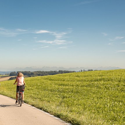 Frau fährt Fahrrad auf Landstraße neben grünem Hügel bei klarem Himmel