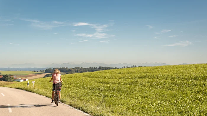 Frau fährt Fahrrad auf Landstraße neben grünem Hügel bei klarem Himmel