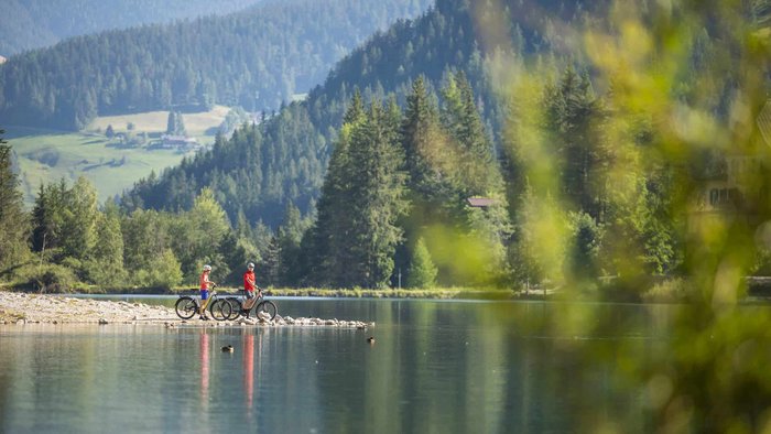 Two people with bicycles by a calm lake with forested mountains