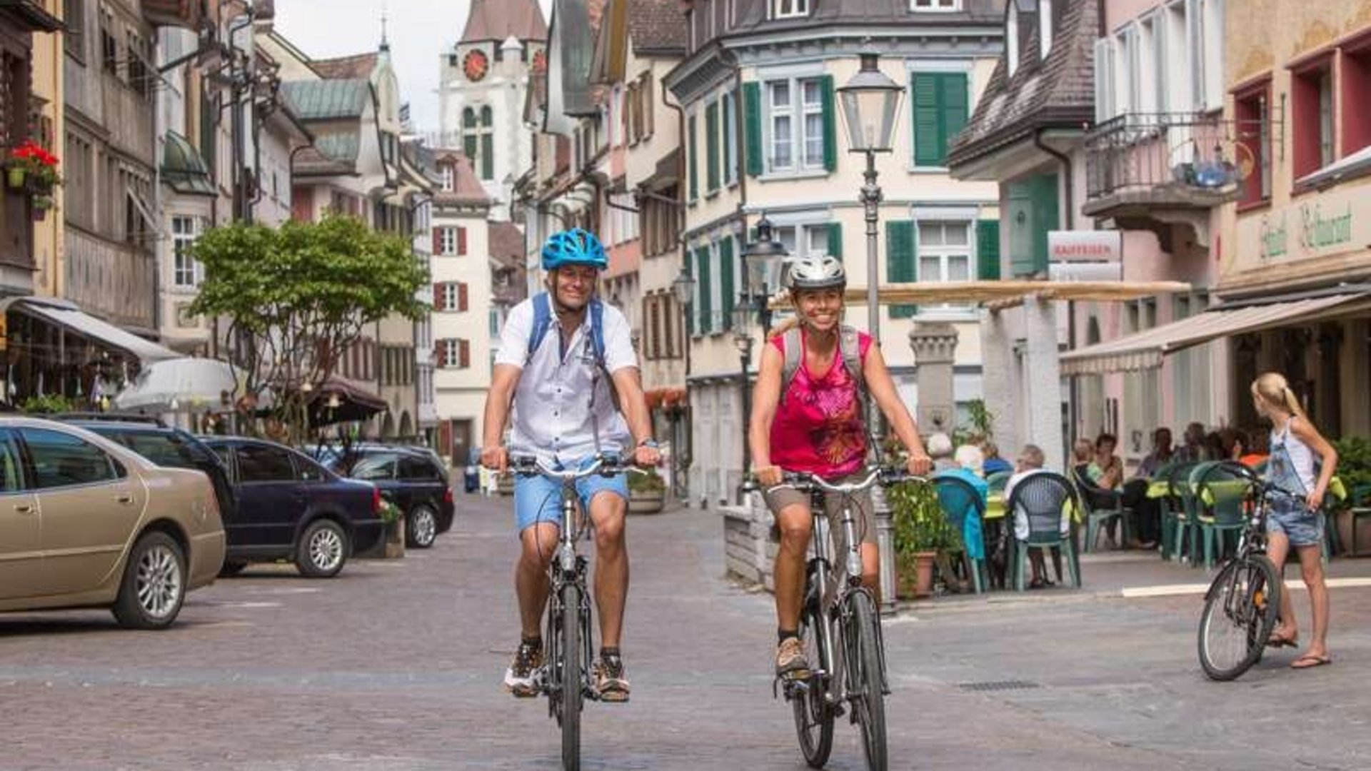 Two people cycling through a picturesque old town with cafes and historic buildings.