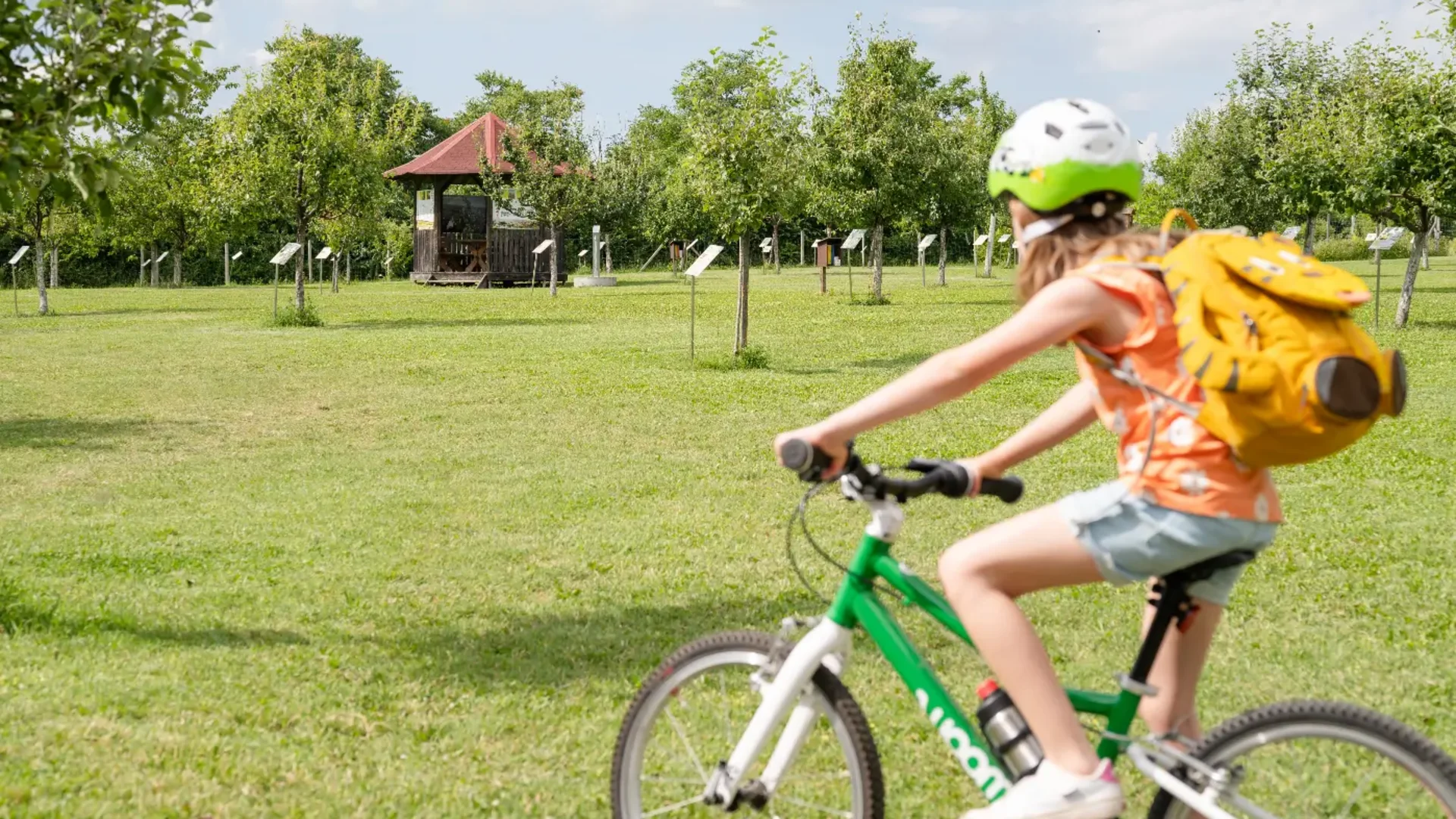 Tour-Detail © Claudia Schlager Kind mit Helm und Rucksack fährt Fahrrad auf grüner Wiese mit Bäumen