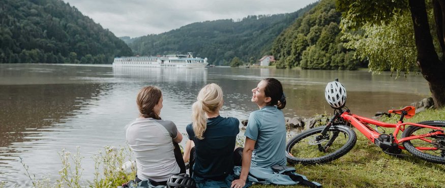 Three women with bikes sitting by river watching a cruise ship