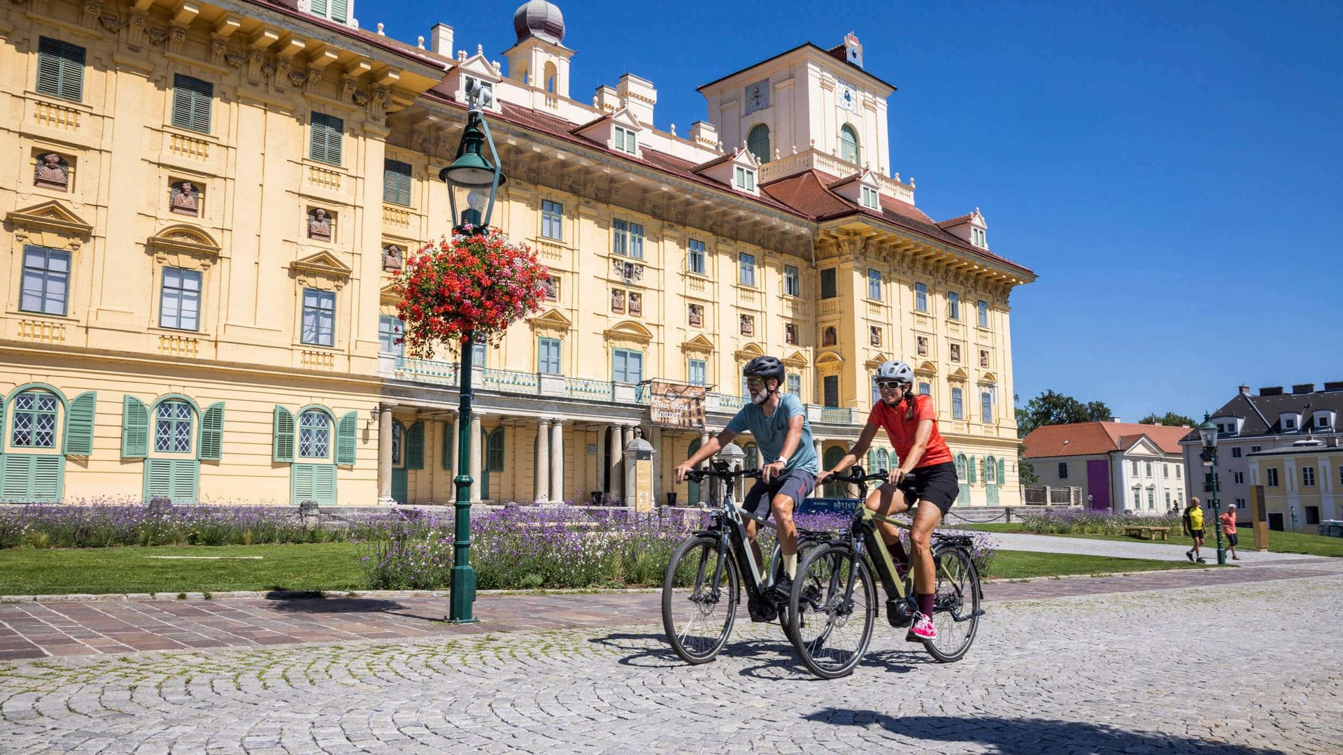 Two people cycling on a sunny day in front of a large yellow palace