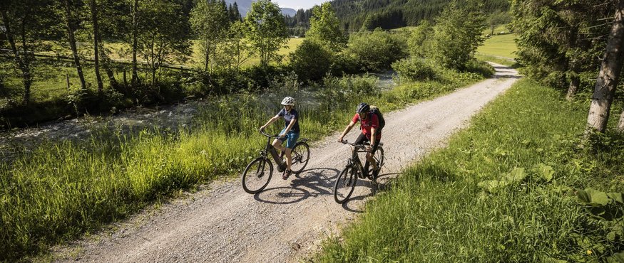 Two cyclists riding on forest path beside stream in mountainous landscape