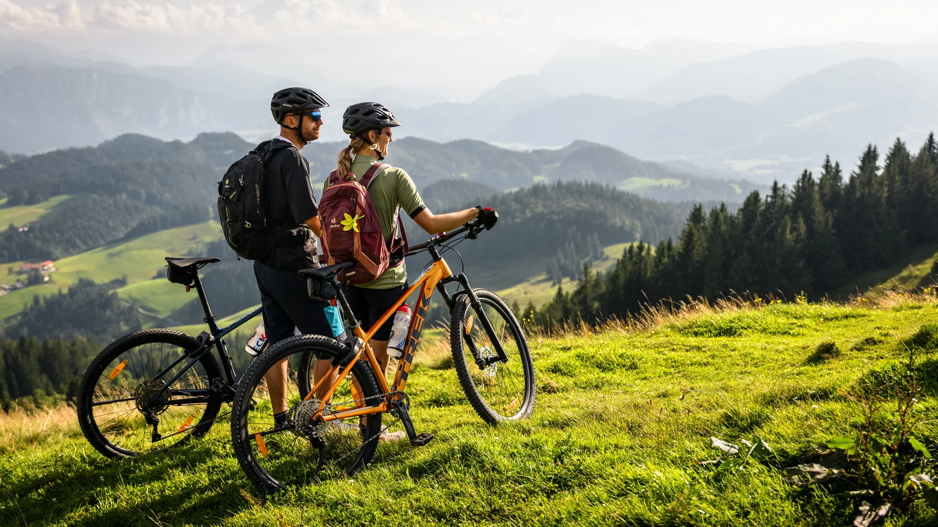Zwei Radfahrer mit Helmen stehen auf einer grünen Wiese und blicken auf Berge