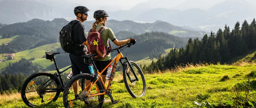 Zwei Radfahrer mit Helmen stehen auf einer grünen Wiese und blicken auf Berge