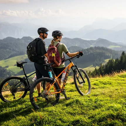 Zwei Radfahrer mit Helmen stehen auf einer grünen Wiese und blicken auf Berge
