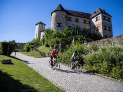Two cyclists riding on cobblestone road with a historic castle in background