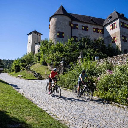 Two cyclists riding on cobblestone road with a historic castle in background