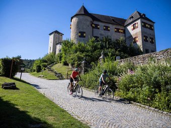 Two cyclists riding on cobblestone road with a historic castle in background