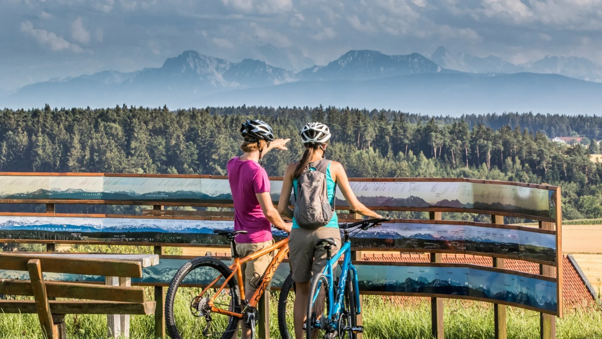 Zwei Radfahrer betrachten eine Panorama-Tafel mit Bergen im Hintergrund