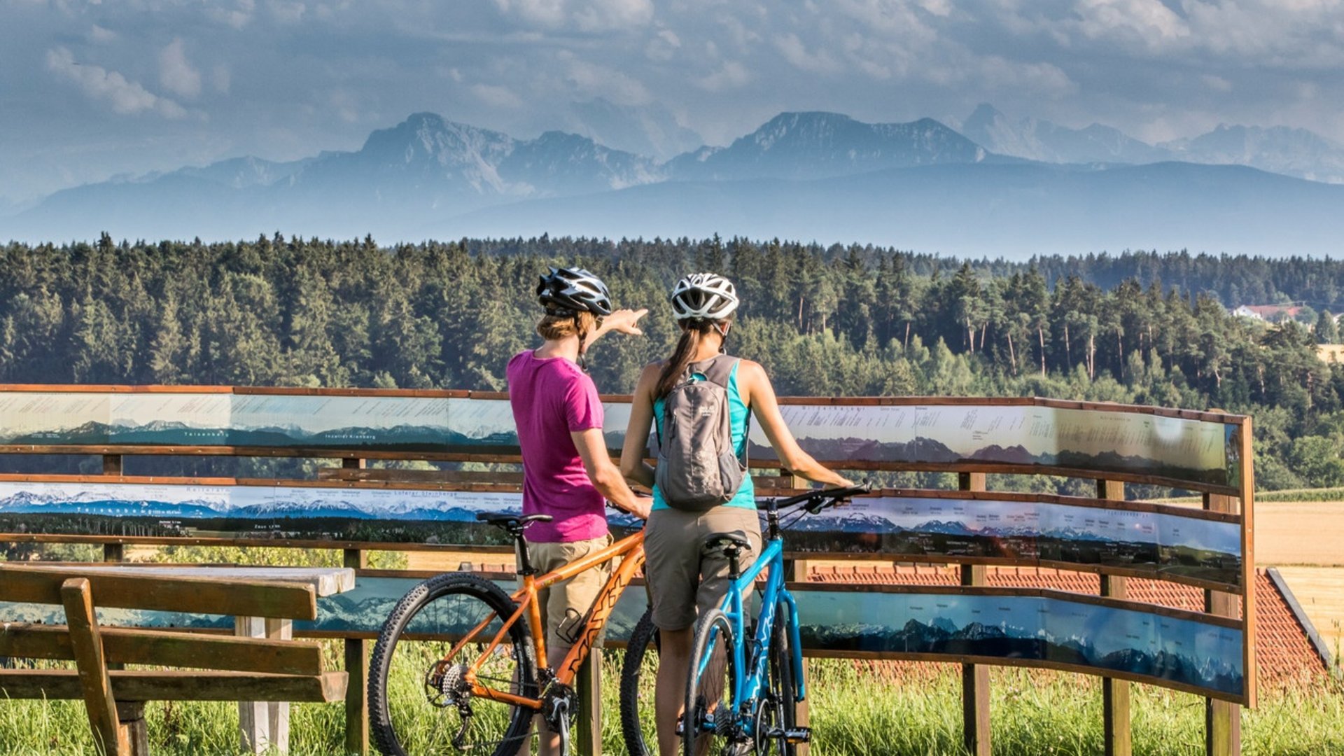 Two cyclists look at a panoramic map with mountains in the background