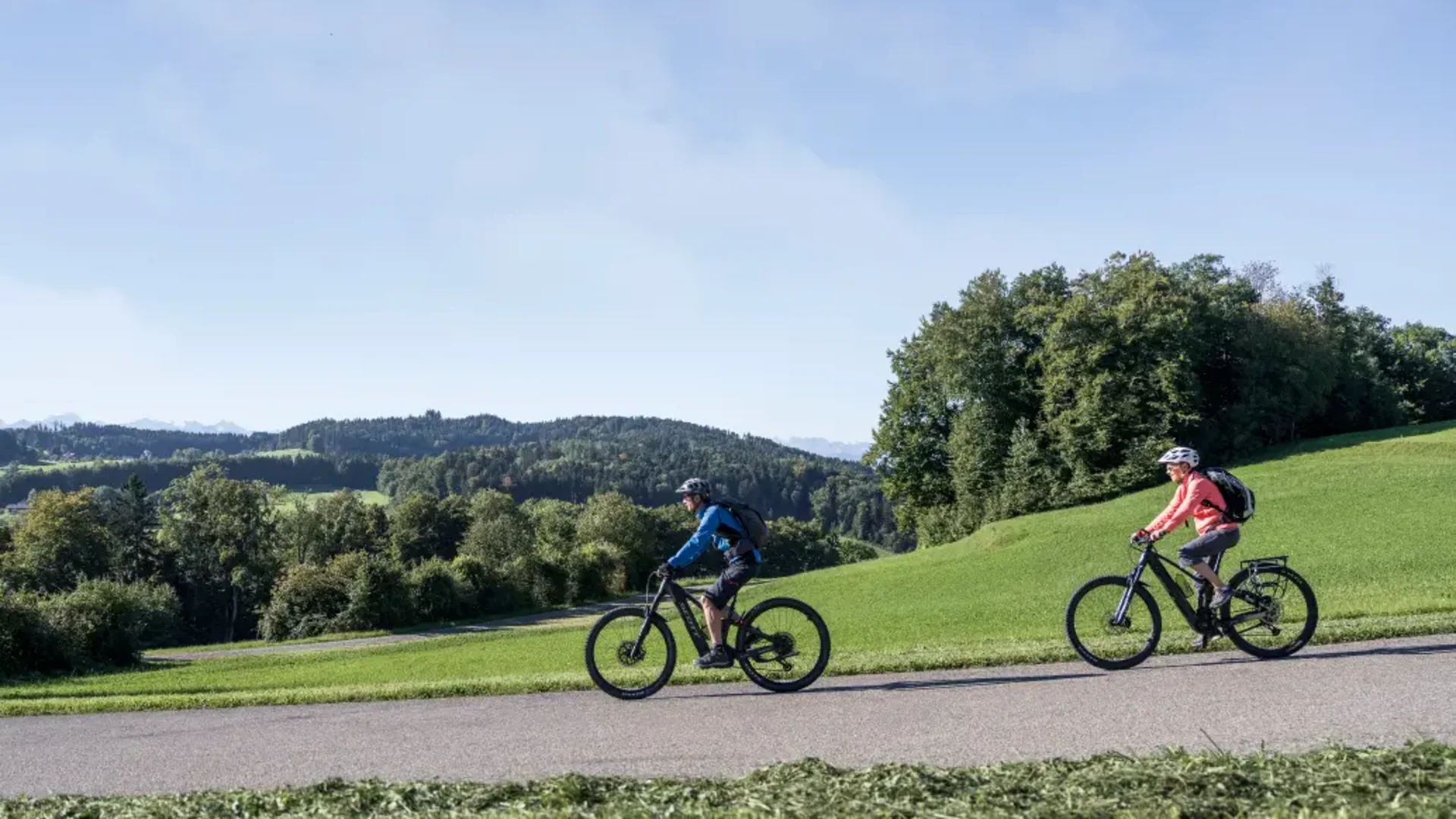Twee mensen fietsen op een weg door een groen heuvelachtig landschap.