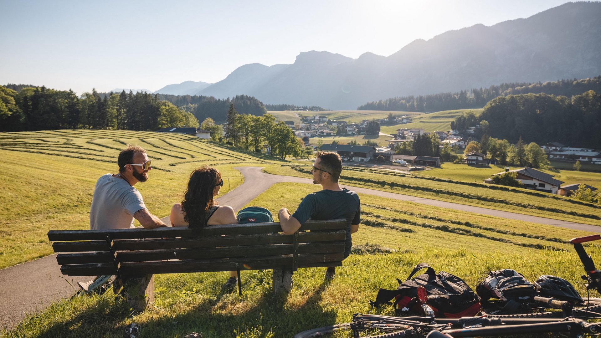 Three people sit on a bench with bikes looking at countryside view