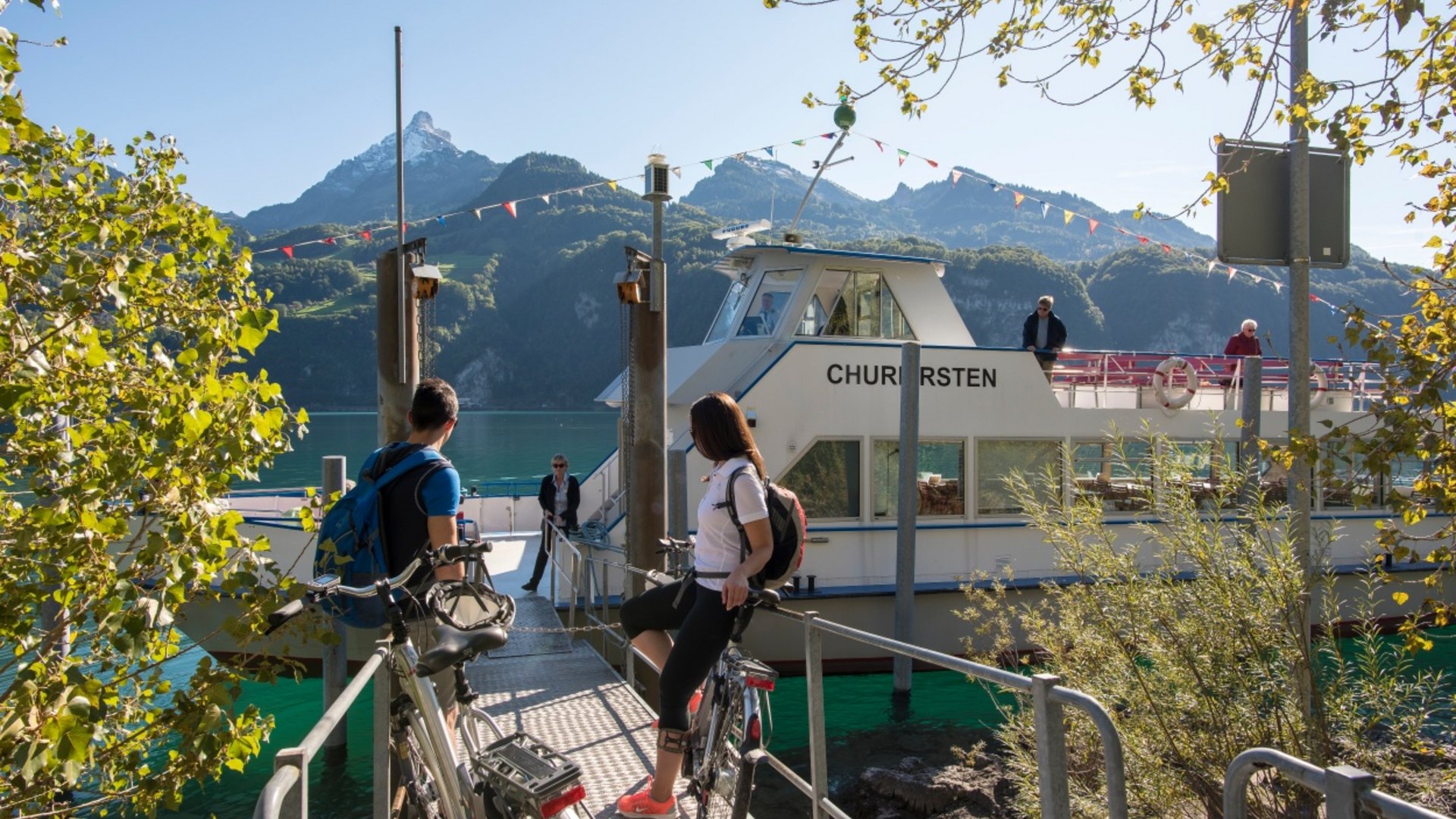 Two cyclists at boat dock with mountains and clear sky in background