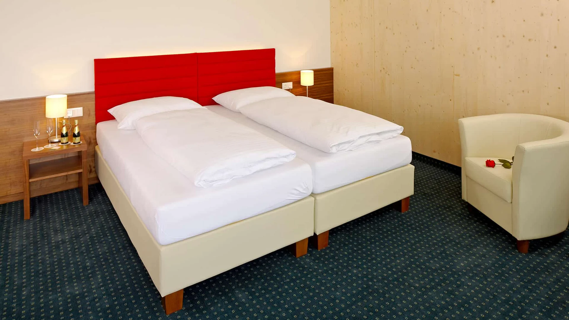 Tweepersoonsbed met rood hoofdeinde, fauteuil en nachtkastje in hotelkamer