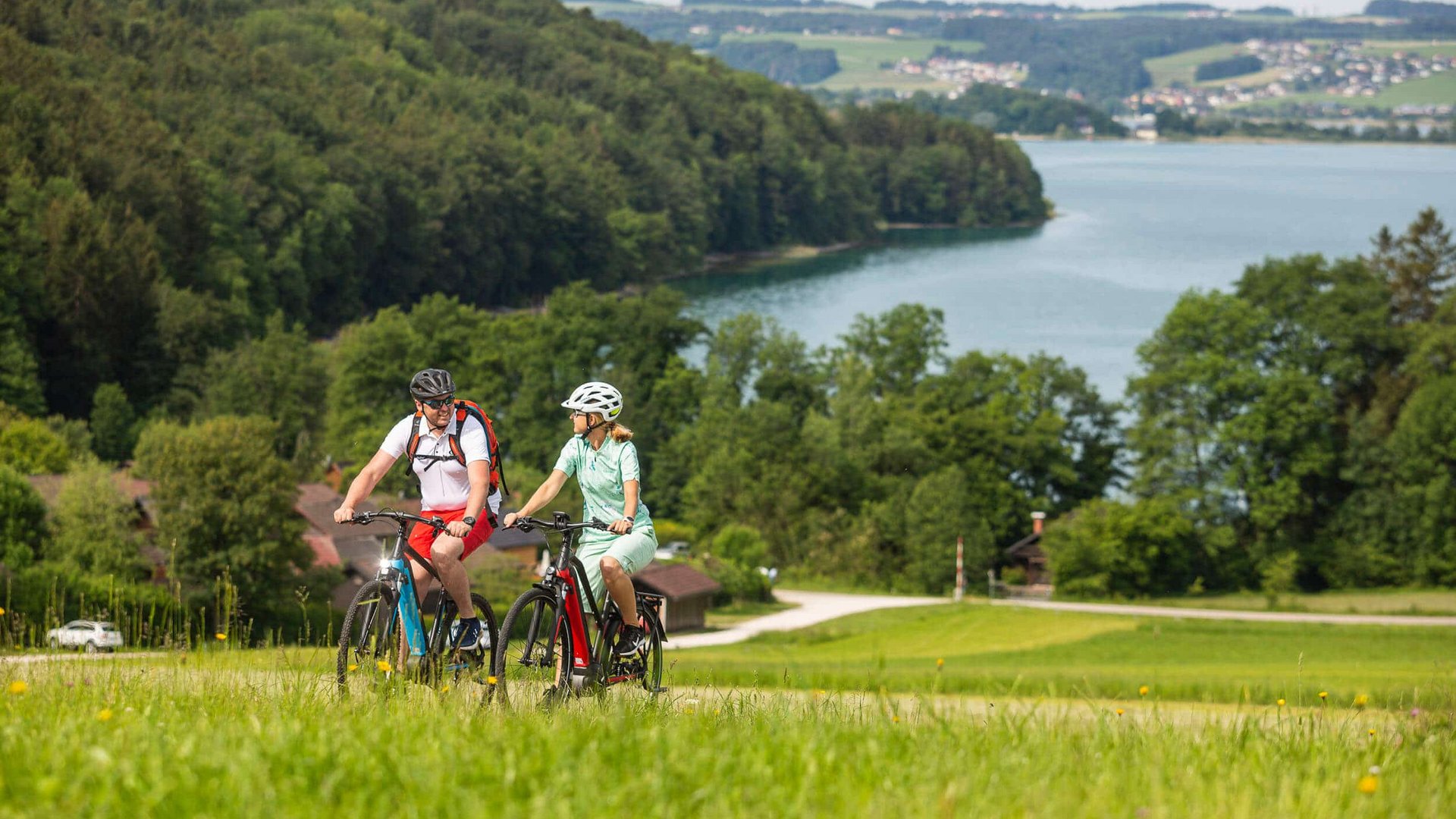 Two cyclists wearing helmets ride on a path in green landscape with lake