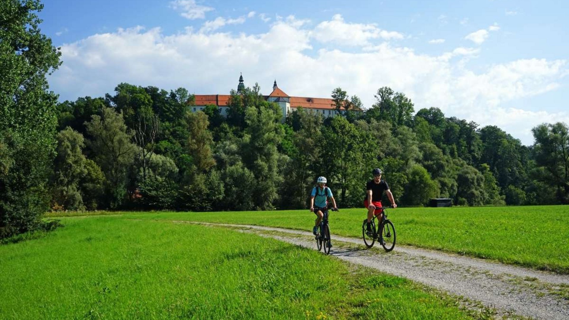 Two cyclists ride on a path in green landscape with a castle on a hill