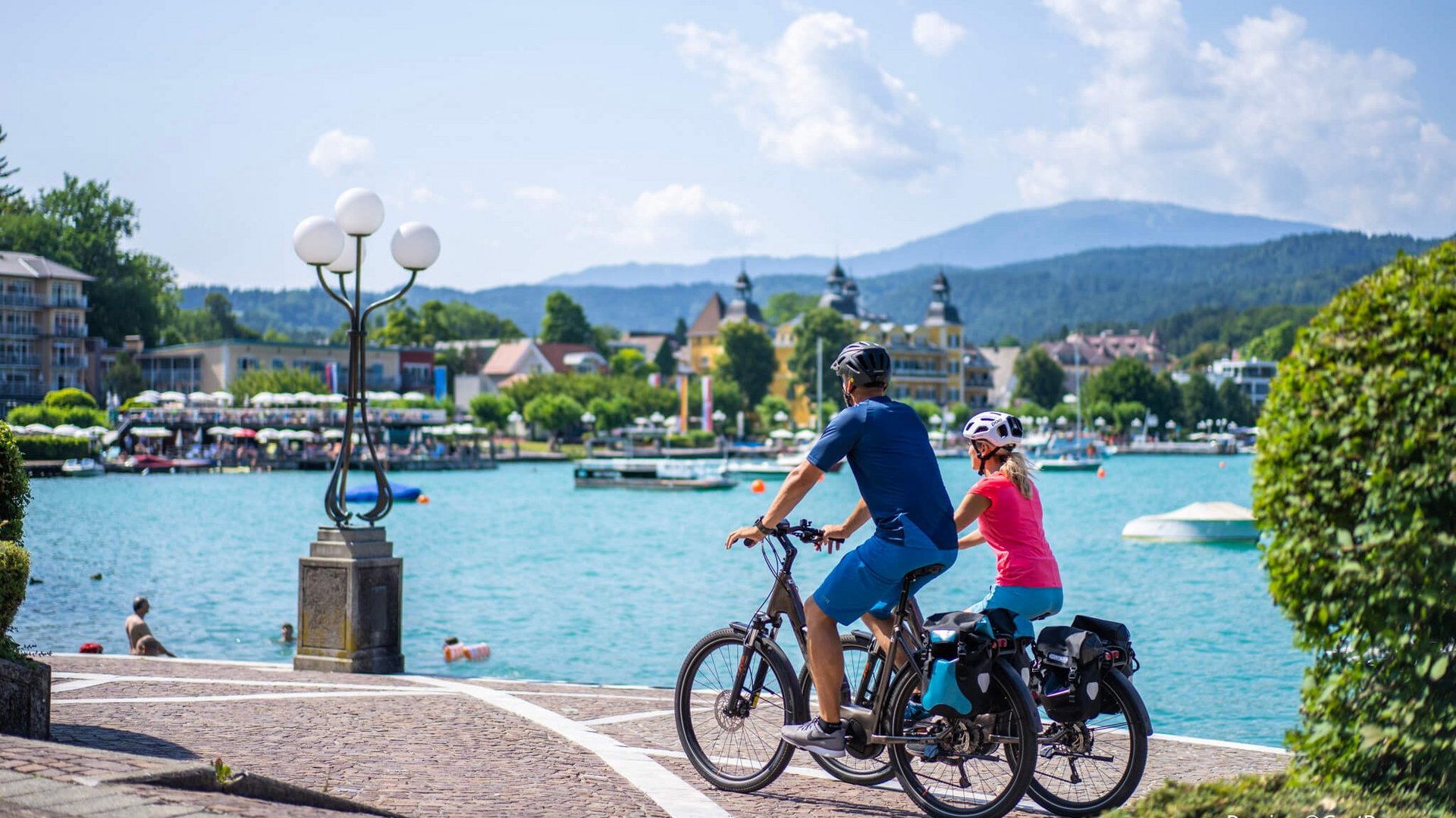 Two cyclists by a lake shore with mountains and buildings in the background