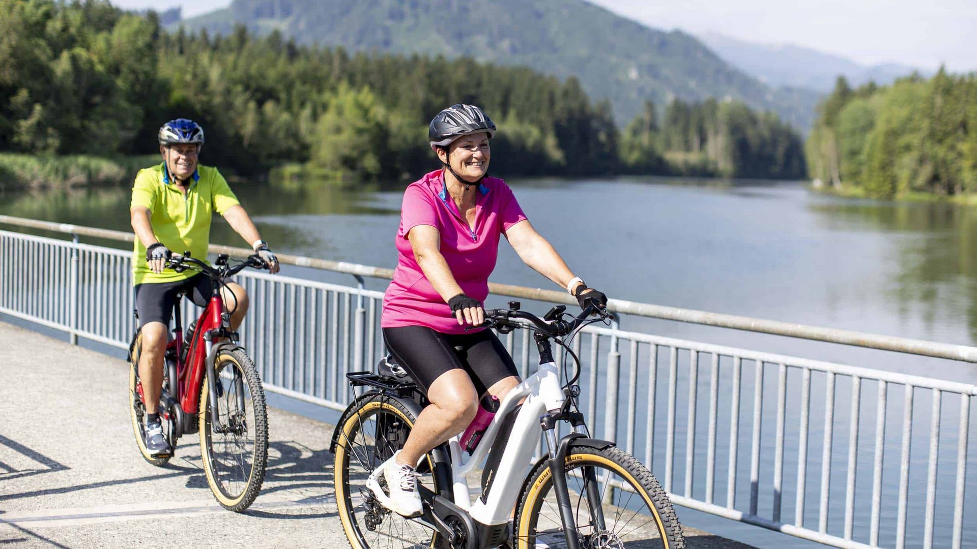 Two seniors cycling on a path by a river with mountains in the background