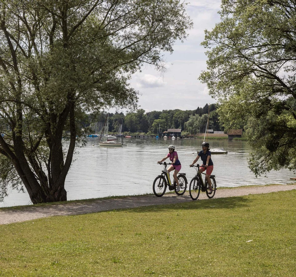 Zwei Radfahrer auf Weg am Seeufer mit Booten und Bänken unter Bäumen