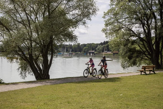 Zwei Radfahrer auf Weg am Seeufer mit Booten und Bänken unter Bäumen