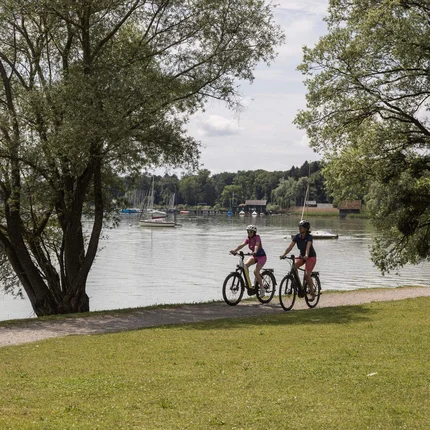 Drei Radwege, drei Seiten Oberbayerns © Velontour Zwei Radfahrer auf Weg am Seeufer mit Booten und Bänken unter Bäumen