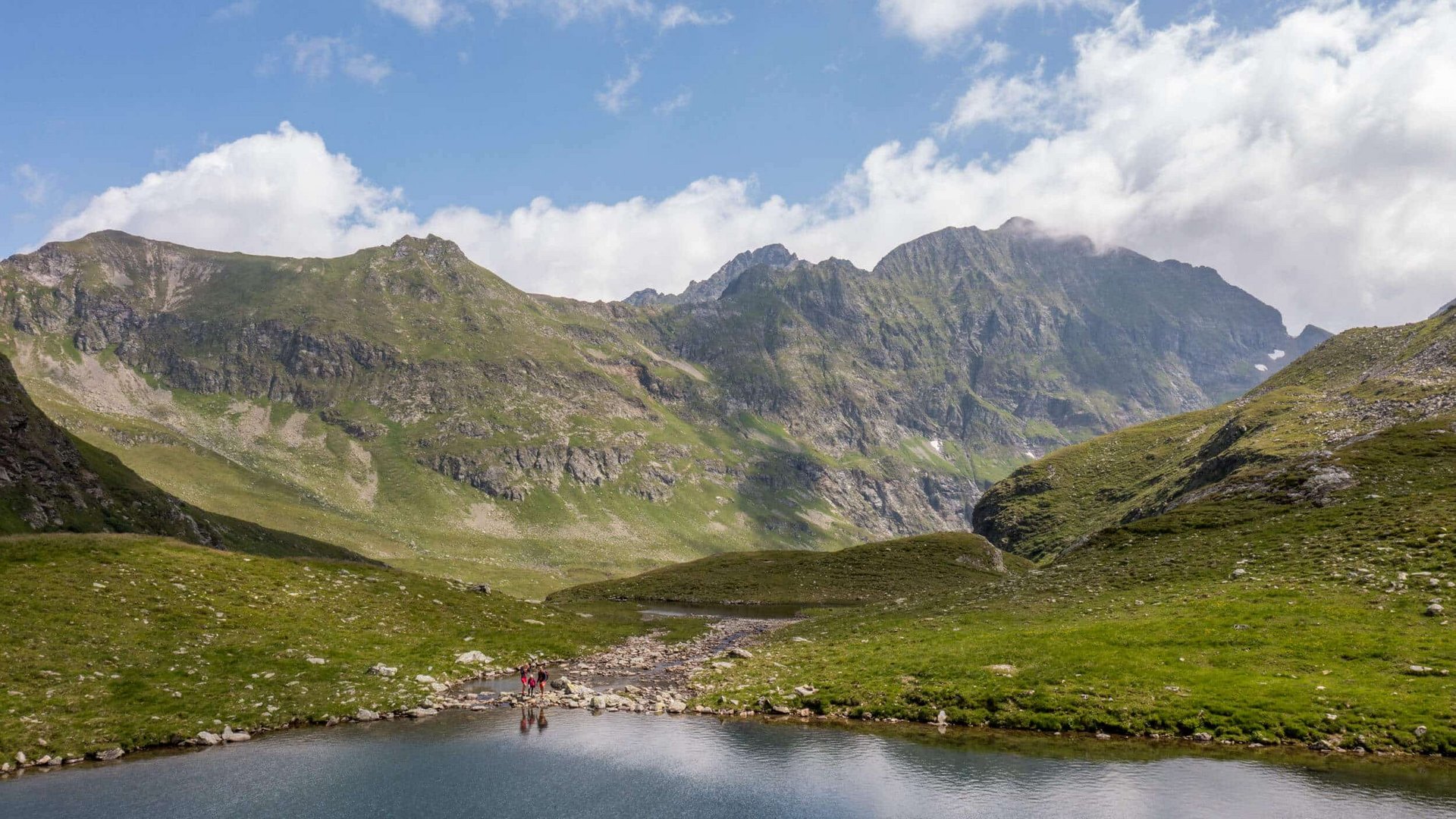 Mountain lake with hikers near green mountains under blue sky