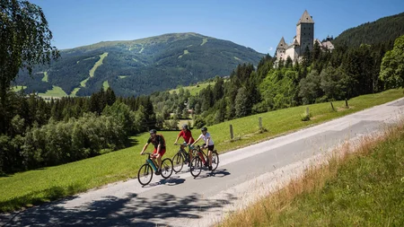Drei Radfahrer fahren auf Landstraße mit Schloss und Bergen im Hintergrund